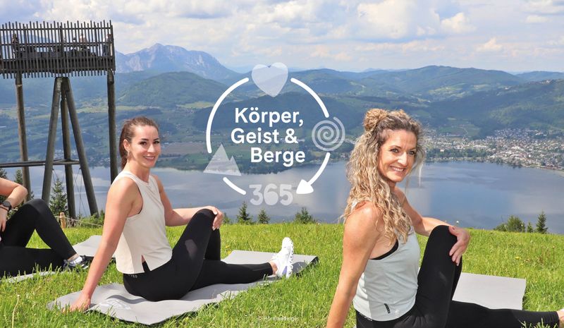 Zwei Frauen sitzen in einer Drehhaltung auf Matten auf einem grasbewachsenen Hügel und lächeln in die Kamera. Malerische Kulisse mit See und Bergen. Text: „Körper, Geist & Berge 365“.