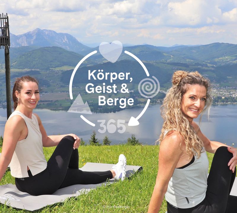 Zwei Frauen sitzen in einer Drehhaltung auf Matten auf einem grasbewachsenen Hügel und lächeln in die Kamera. Malerische Kulisse mit See und Bergen. Text: „Körper, Geist & Berge 365“.