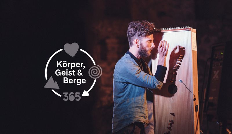 Ein bärtiger Mann spielt ein großes, stehendes Saiteninstrument auf einer dunklen Bühne. Links ist das Logo "Körper, Geist & Berge 365" zu sehen.