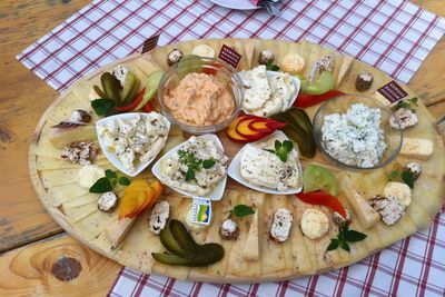 An oval wooden board laden with various cheeses, dips, pickles, and herbs on a checkered tablecloth.