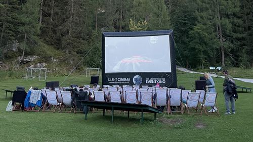 Ein Freiluftkino mit großer Leinwand und Reihen von Liegestühlen auf einer grünen Wiese, umgeben von Wald und Felswänden unter blauem Himmel.