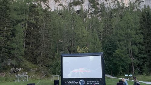 An open-air cinema with a large screen and rows of deckchairs on a green meadow, surrounded by forest and rock faces under a blue sky.