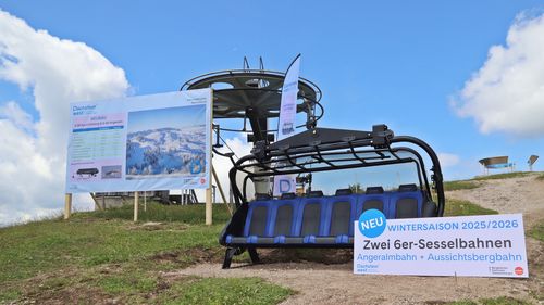 Sessellift mit blauen Sitzen im Freien auf einer Wiese, mit Werbeschildern und einem bewölkten Himmel im Hintergrund.