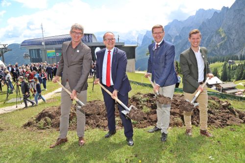Four men in suits smile and hold shovels at a ground-breaking ceremony with mountains and a crowd of people in the background