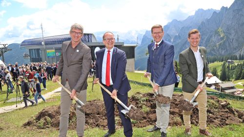 Four men in suits smile and hold shovels at a ground-breaking ceremony with mountains and a crowd of people in the background