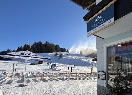 Verschneites Skigebiet mit klarem blauen Himmel, einem Gebäude rechts und schneebedeckten Hügeln im Hintergrund. Menschen und Skiausrüstung sichtbar.
