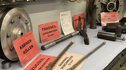 Seilbahnteile auf einem Tisch: Trag-, Zug- und Gegenseile mit verschiedenen Durchmessern und Kabinenrollen, beschriftet mit roten Karten.