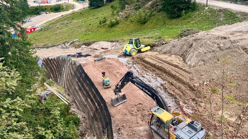 Baustelle in einer bergigen Gegend mit Baggern und Arbeitern auf einem Erdhang, umgeben von üppig grünen Bäumen und Hügeln.