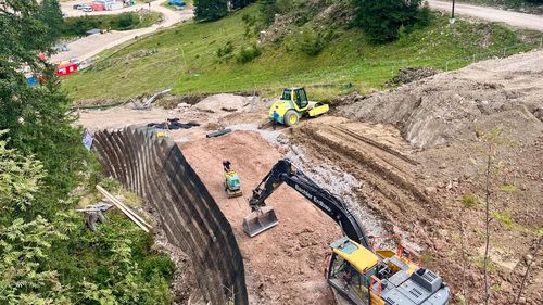 Construction site in a mountainous area with excavators and workers on a slope, surrounded by lush green trees and hills.
