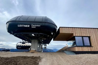Eine moderne Seilbahnstation mit einem Holzgebäude und einer Gondel mit der Aufschrift „LEITNER Dachstein West” vor einem bewölkten Himmel.