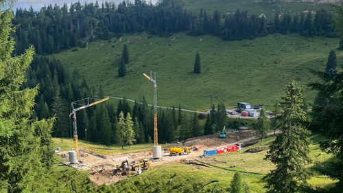Construction site in a lush green valley with cranes, machinery and surrounding trees under a partly cloudy sky.