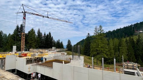 Baustelle in einer hügeligen Landschaft mit Kränen, Baumaschinen und strahlend blauem Himmel. Das Gebiet ist von bewaldeten Hügeln umgeben.