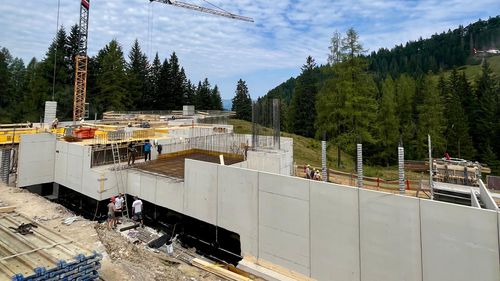 Construction site with crane, workers and partially completed concrete structures against a backdrop of trees and cloudy sky.