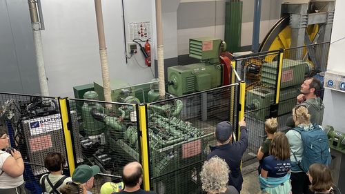 Eine Gruppe Menschen besichtigt einen Maschinenraum mit großen grünen Maschinen und einem gelben Rad, während ein Mann etwas erklärt.