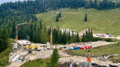 Construction site in hilly landscape with cranes, construction machinery and bright blue sky. The area is surrounded by wooded hills.