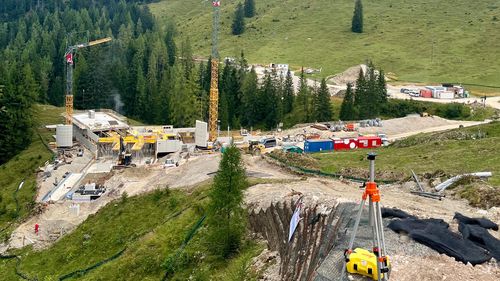 Baustelle in einer hügeligen Landschaft mit Kränen, Baumaschinen und strahlend blauem Himmel. Das Gebiet ist von bewaldeten Hügeln umgeben.