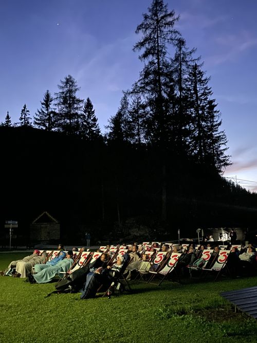 At dusk, audience members sit in deckchairs on a lawn in front of a dark forest backdrop at an open-air cinema.