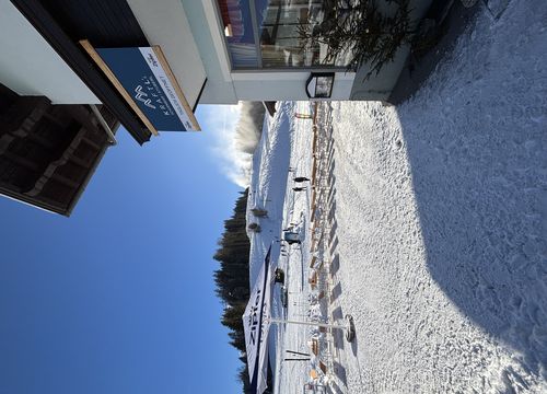 Snowy ski resort with clear blue sky, a building on the right and snow-covered hills in the background. People and ski equipment visible.