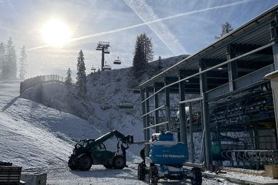 Snowy construction site on a mountain with machinery, metal beams, and a ski lift in the background under a bright sun and clear sky.