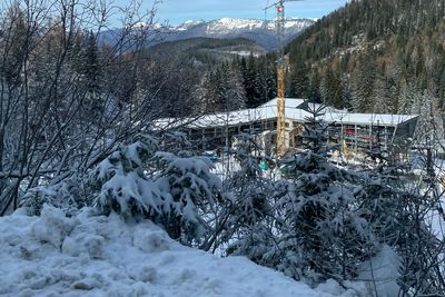 Snow-covered landscape with evergreen trees, a construction crane, and mountains in the background under a blue sky.