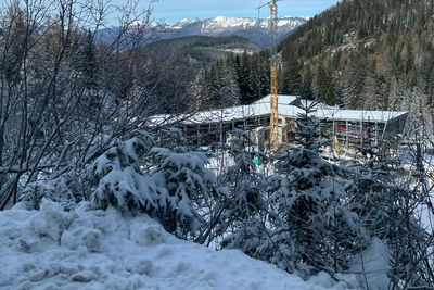 Schneebedeckte Landschaft mit immergrünen Bäumen, einem Baukran und Bergen im Hintergrund unter blauem Himmel.