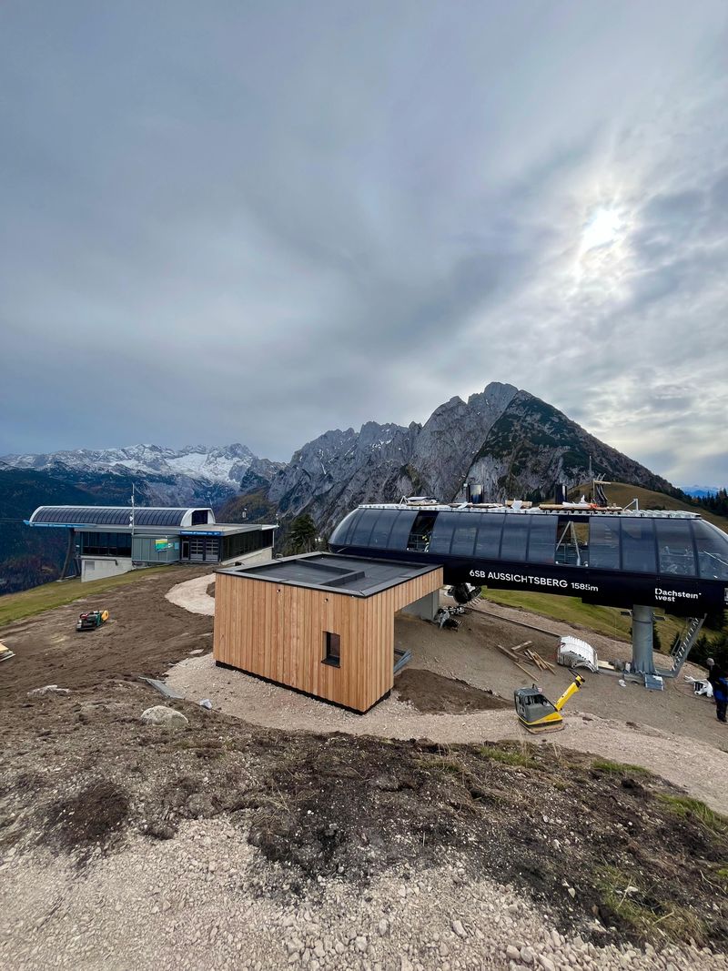 Mountain cable car station with wooden building, rugged peaks in the background, and a cloudy sky above. Dirt paths surround the area.