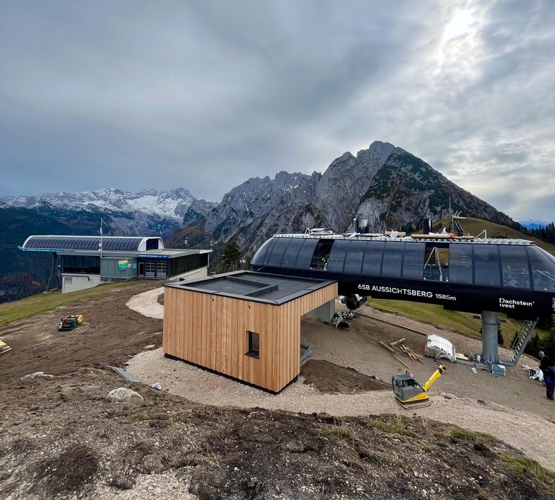 Bergbahnstation mit Holzgebäude, schroffen Gipfeln im Hintergrund und bewölktem Himmel darüber. Feldwege umgeben das Gebiet.
