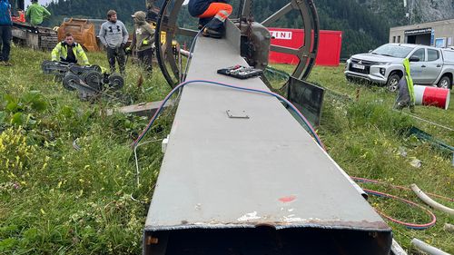 Workers use machinery to dismantle a large metal structure in a mountainous landscape. An excavator and vehicles can be seen in the background.