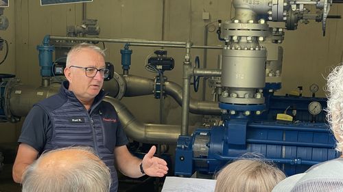 Ein Mann erklärt einer Gruppe die Beschneiungsanlage vor Rohren, Maschinen und Fotos von Winterlandschaften mit Schneekanonen.