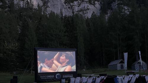 "Silent Cinema" open-air evening movie event: a large screen shows a film, with empty deckchairs on the lawn in front of a wooded, rocky hill.