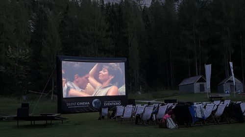 "Silent Cinema" Eventkino im Freien am Abend: Eine Großleinwand zeigt einen Film, leere Liegestühle auf der Wiese vor bewaldetem Felsberg.