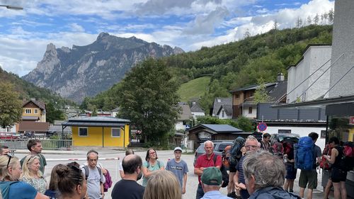 Gruppe Wanderer mit Rucksäcken in einem Dorf mit bewaldeten Bergen und wolkigem Himmel im Hintergrund.