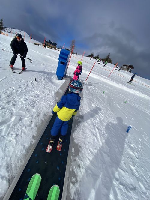 Kinder mit Skihelm fahren im Drachenpark auf einem Förderband bergauf, begleitet von Erwachsenen im Schnee.
