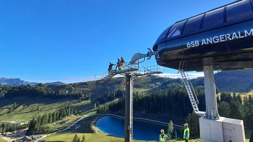 Arbeiter bei der Installation einer Seilbahnstütze für eine Sesselbahn mit malerischem Blick auf Berge, einem Speicherteich und einen strahlend blauen Himmel
