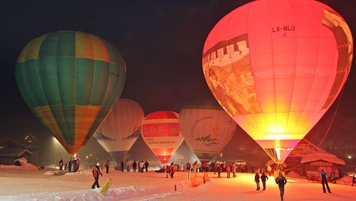 Nachts beleuchtete Heißluftballons, die auf der verschneiten Skipiste stehen, um die sich Menschen versammelt haben, schaffen eine lebendige und festliche Atmosphäre.