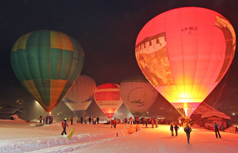 At night, illuminated hot-air balloons placed on the snow-covered ski slope, around which people have gathered, create a lively and festive atmosphere.