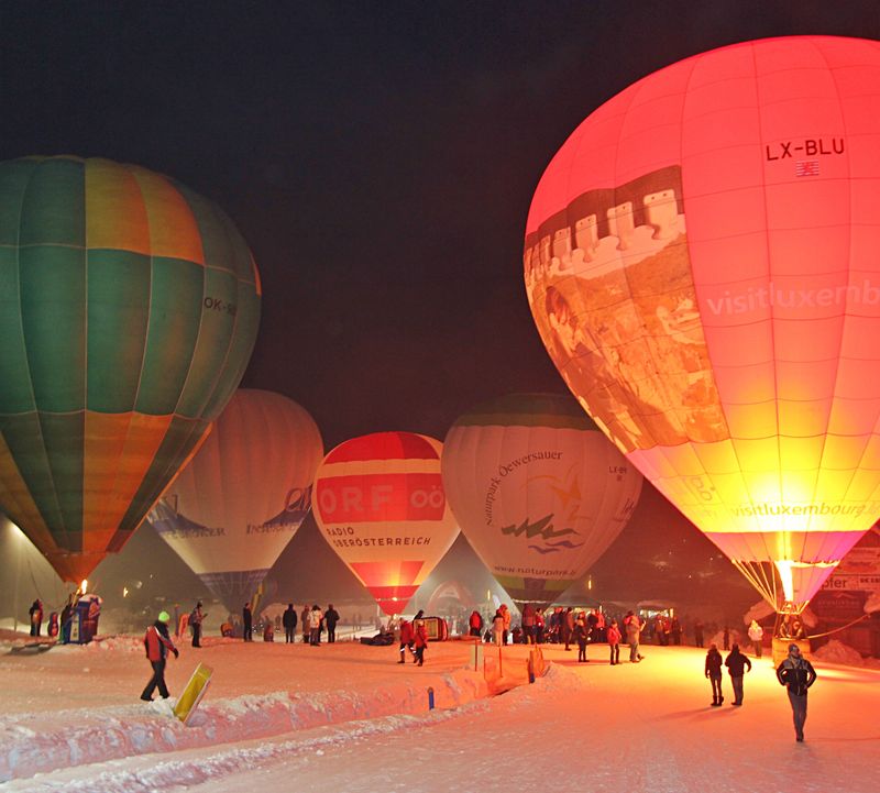 Nachts beleuchtete Heißluftballons, die auf der verschneiten Skipiste stehen, um die sich Menschen versammelt haben, schaffen eine lebendige und festliche Atmosphäre.