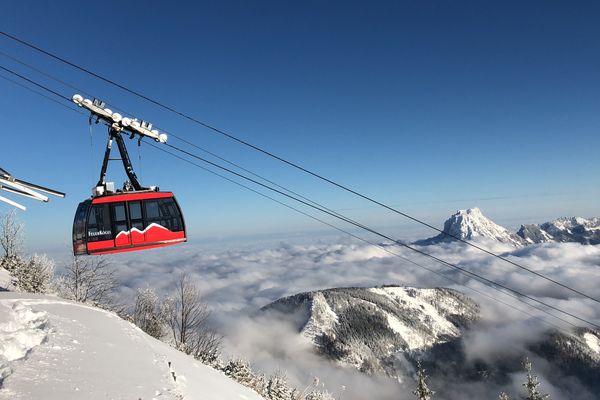Eine rote Feuerkogel-Gondel fährt über das verschneite Tal, im Hintergrund ragt der Dachstein über ein weißes Wolkenmeer.