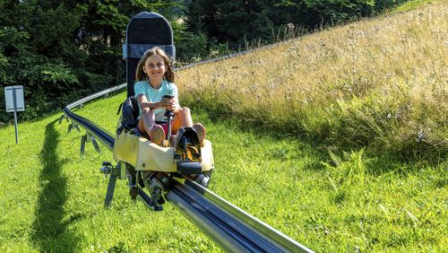 Person fährt mit gelbem Sommerrodelbahn-Wagen eine grüne Hügelwiese hinunter, umgeben von Bäumen und strahlendem Sonnenschein.