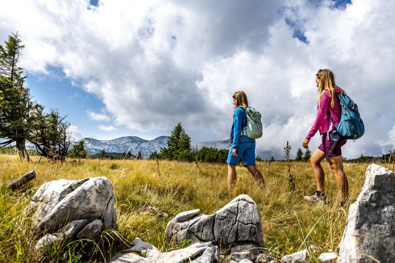 Two women with rucksacks hike through an open meadow landscape with a view of cloud-covered mountains.