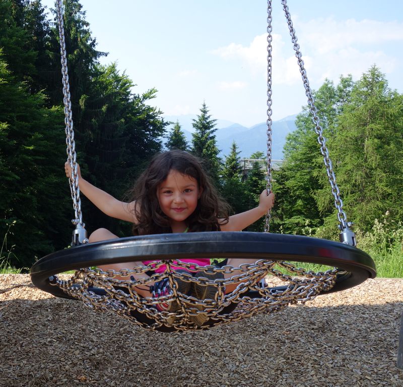 Mädchen mit langen braunen Haaren sitzt auf einer runden Schaukel, umgeben von Bäumen, Berge im Hintergrund bei sonnigem Himmel.