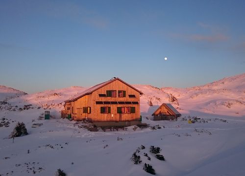 The Rieder Hütte and a smaller building stand in a snowy landscape at sunset, with a clear sky above and the moon visible.