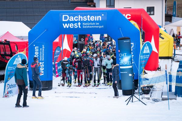 Die Skifahrer versammelten sich an einer verschneiten Startlinie unter einem blauen Torbogen mit der Aufschrift „Dachstein West“, umgeben von bunten Zelten und Zuschauern.
