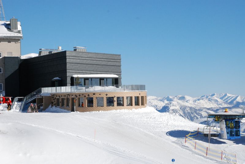Modern mountain station with restaurant in the snow, snow-covered mountain peaks and blue sky behind it.