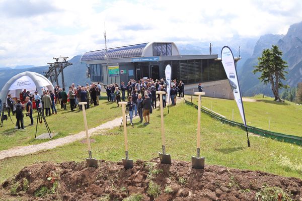 Eine Gruppe von Menschen versammelt sich in der Nähe eines modernen Seilbahngebäudes auf einem grasbewachsenen Hügel mit im Boden steckenden Schaufeln, umgeben von Bergen und einem bewölkten Himmel.