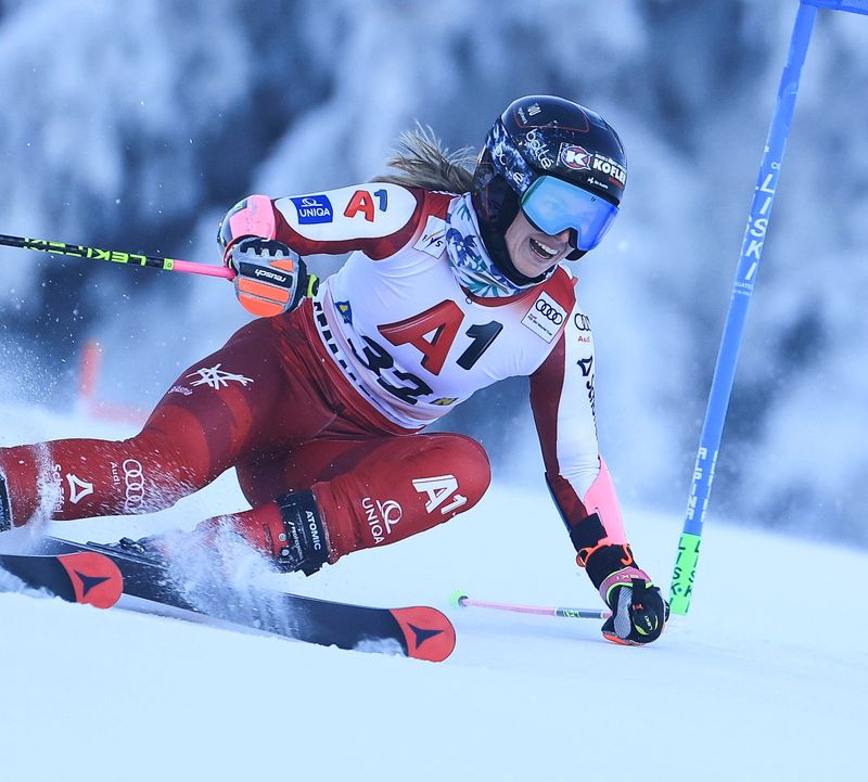 A female ski racer in a red racing suit skis through a gate in a deep, banked turn while snow kicks up.
