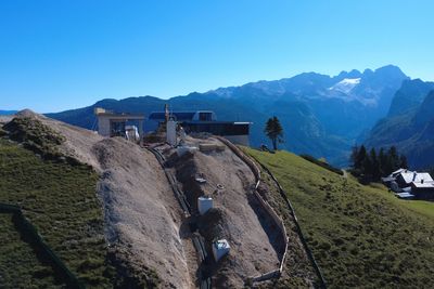Mountain landscape with a building under construction on a grassy slope, surrounded by mountains and a clear blue sky.