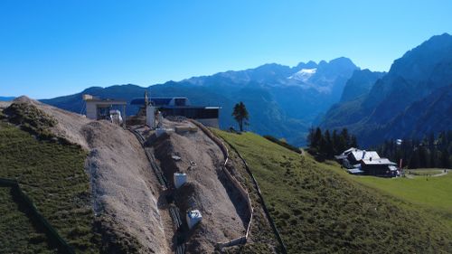 Berglandschaft mit einem im Bau befindlichen Gebäude auf einem grasbewachsenen Hang, umgeben von Bergen und einem klaren blauen Himmel.
