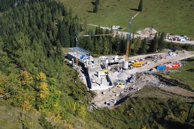 Aerial view of a construction site in a forest area with cranes, machinery and partially completed buildings, surrounded by trees.