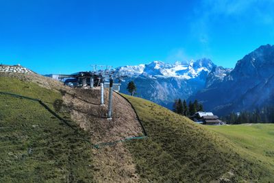 Berglandschaft mit einem Skilift auf einem grasbewachsenen Hang, einer Hütte und schneebedeckten Gipfeln unter strahlend blauem Himmel.